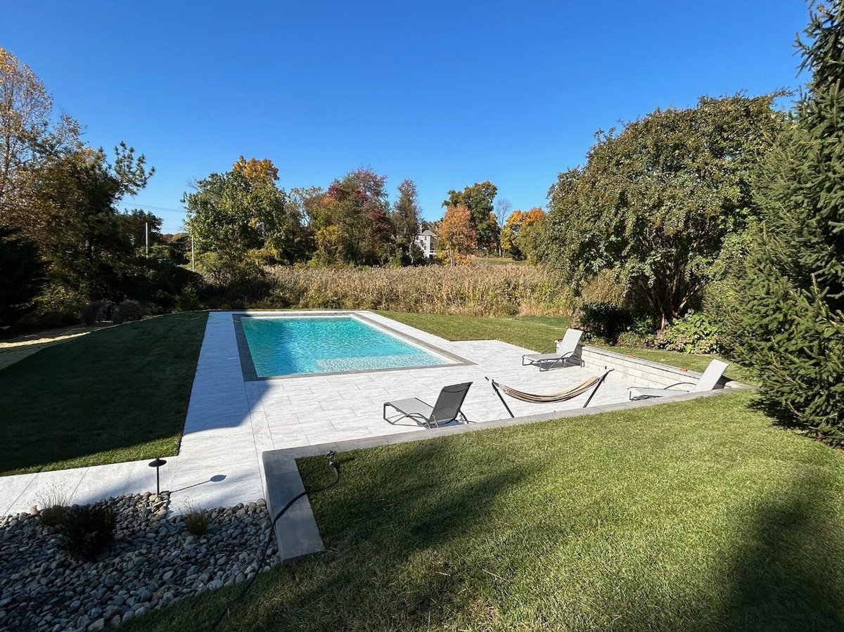 A luxurious backyard with a custom in-ground pool, porcelain patio, and lounge area surrounded by greenery in Newark, DE by First Class Lawn Care.