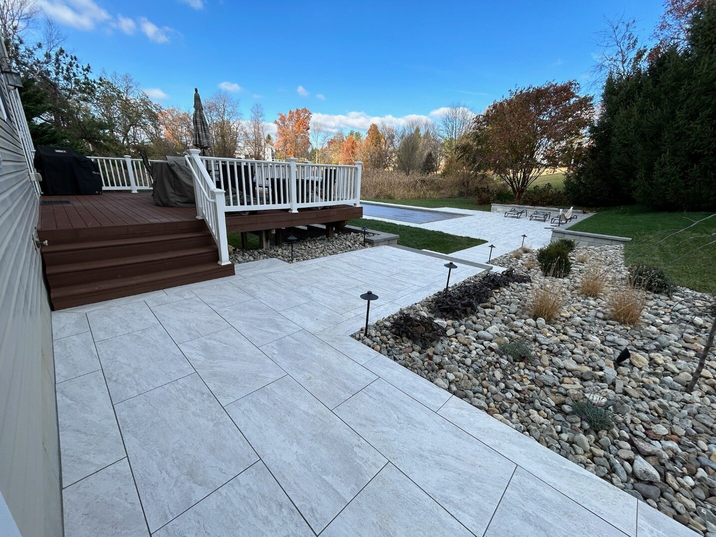 A spacious backyard patio with porcelain pavers, a wooden deck, and a stunning pool in Newark, DE by First Class Lawncare LLC